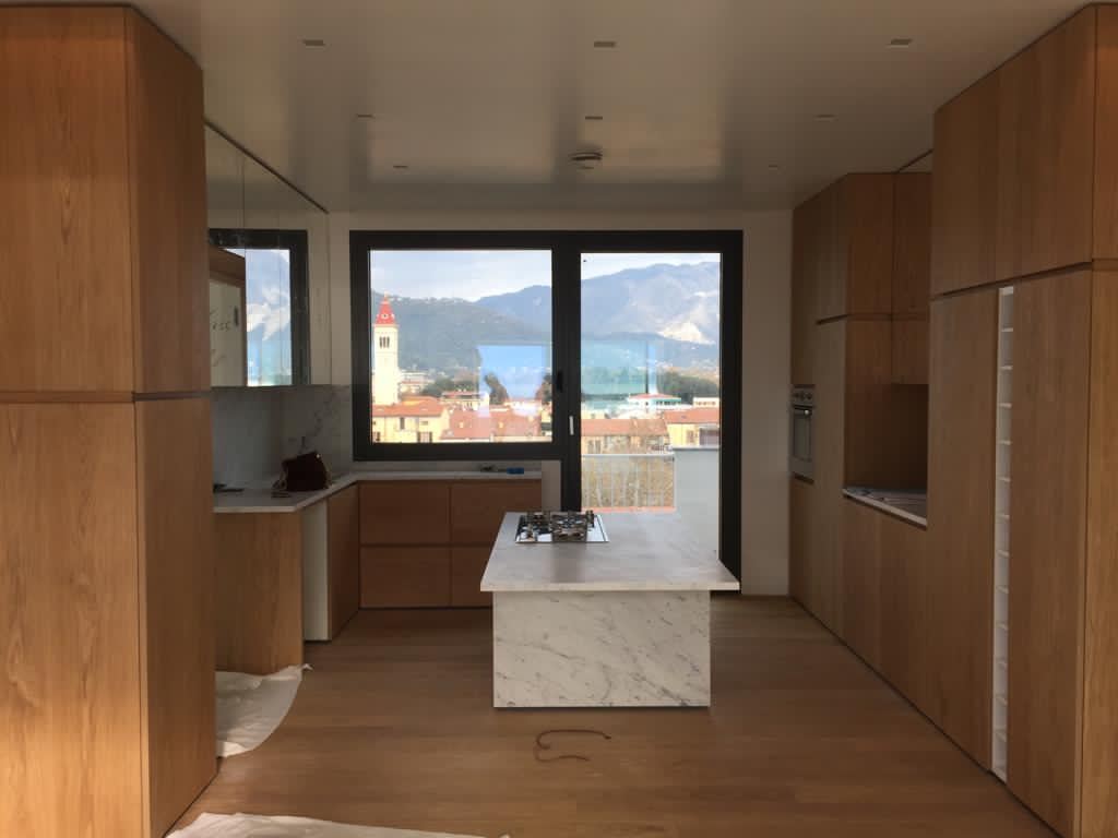 La cucina finita: colonne in rovere ai lati, l'isola in marmo di Carrara al centro, e oltre la vetrata i tetti, il campanile e il mare