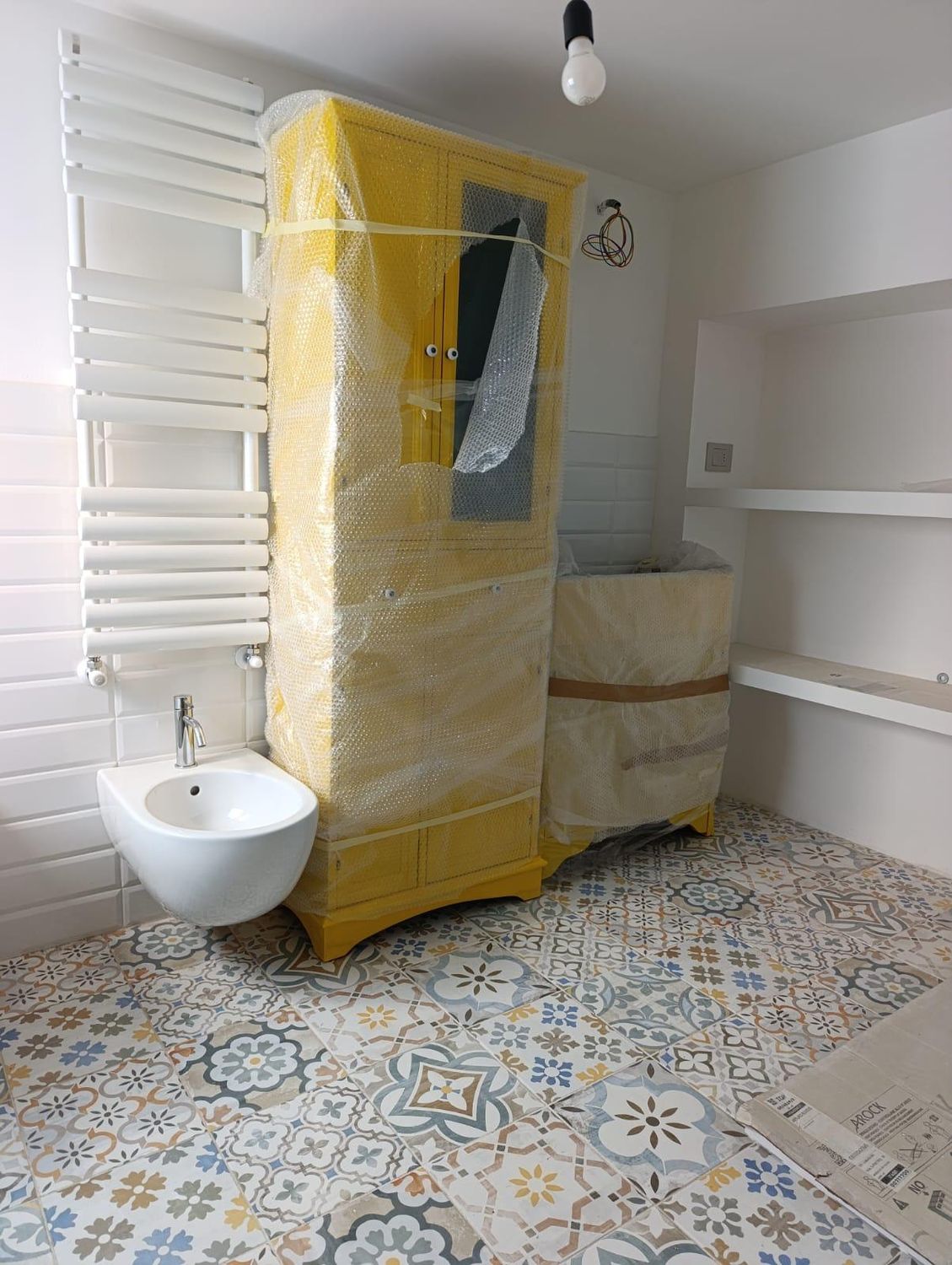 Il bagno piccolo: pavimento a cementine decorate con disegni diversi nei toni dell'azzurro e dell'ocra, pareti piastrellate di bianco, bidet sospeso, termoarredo e mobile ancora avvolto nel pluriball