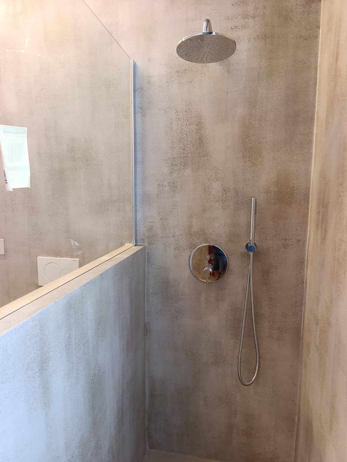 La doccia del bagno padronale: soffione a pioggia, miscelatore e doccetta su parete in resina effetto cemento, con il parapetto in muratura e il vetro fisso