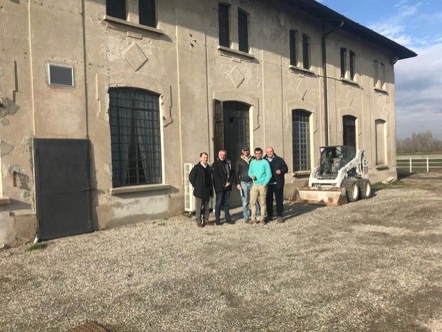 Cinque persone in piedi davanti al fronte lungo del casolare, con una minipala Bobcat accanto