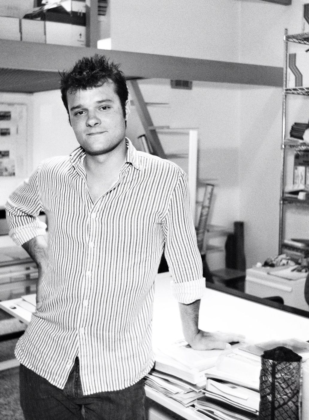 Ritratto in bianco e nero: Matteo Gardino in piedi al tavolo da disegno dello studio, una mano sul fianco e l&rsquo;altra appoggiata sui fogli, gli scaffali metallici alle spalle