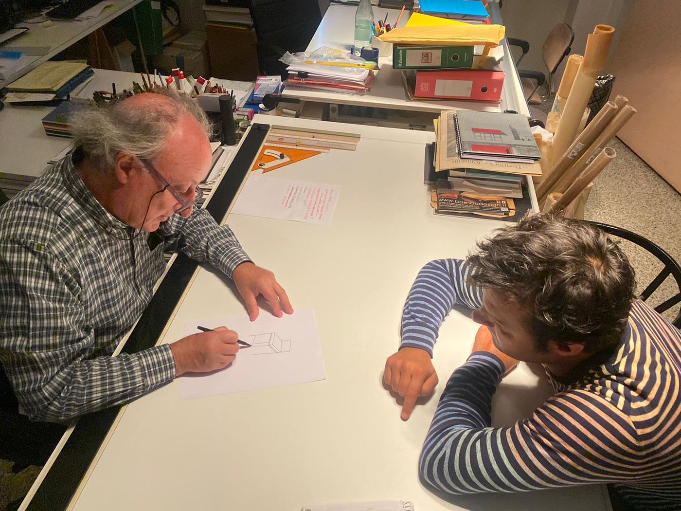 Alberto Gardino disegna a mano un mobile in assonometria sul tavolo da disegno dello studio, Matteo Gardino seduto di fronte indica un punto del foglio