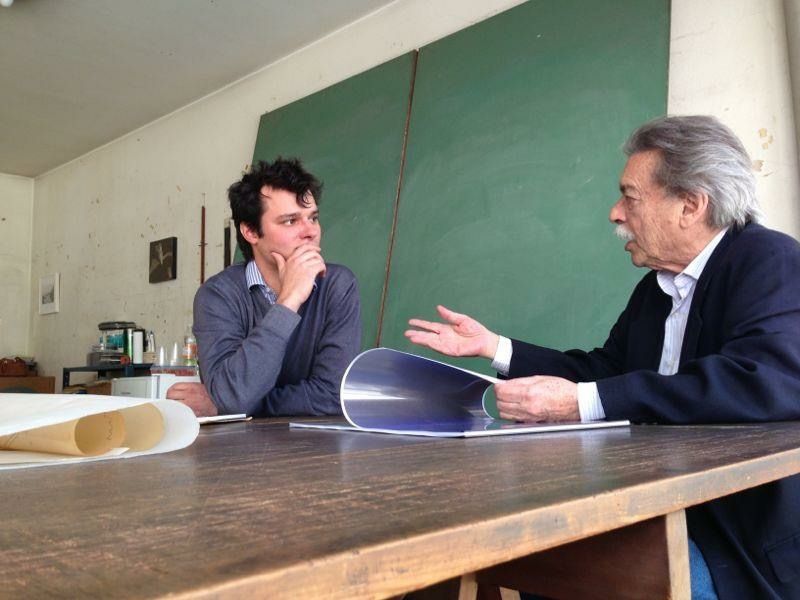 Nello studio di Paulo Mendes da Rocha a San Paolo: lui sfoglia un album di disegni seduto al tavolo di legno, Matteo Gardino ascolta, dietro una lavagna verde