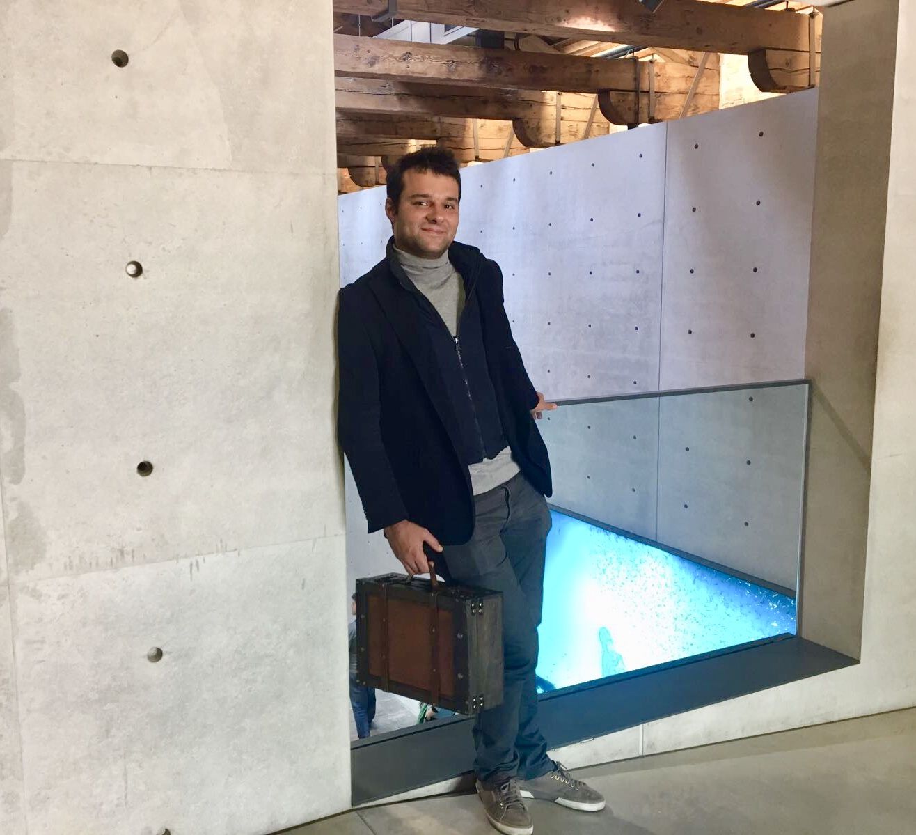Matteo Gardino con una valigia in mano, in piedi dentro un varco aperto in un muro di cemento a vista, sotto le capriate di legno della vecchia dogana e sopra un pavimento di vetro illuminato