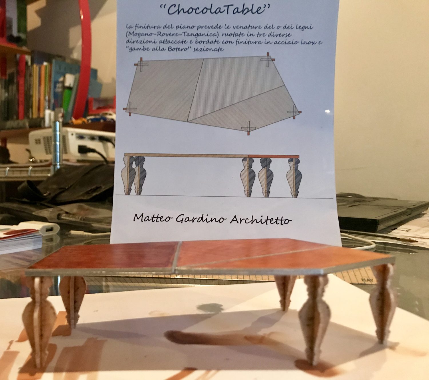 Il modellino davanti al pannello di presentazione in piedi, su cui si leggono il nome ChocolaTable, la nota sui tre legni con le venature ruotate e la dicitura gambe alla Botero sezionate