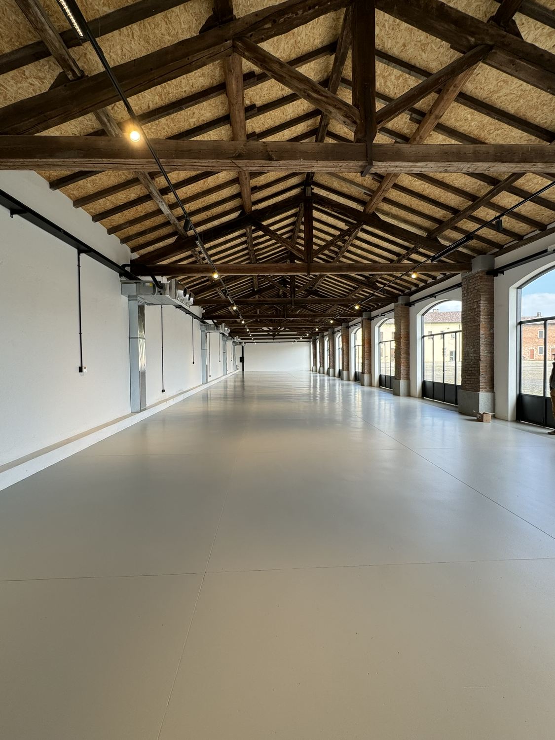 Fotografia della sala lunga finita e vuota: pavimento chiaro, capriate in legno, arcate sul lato destro