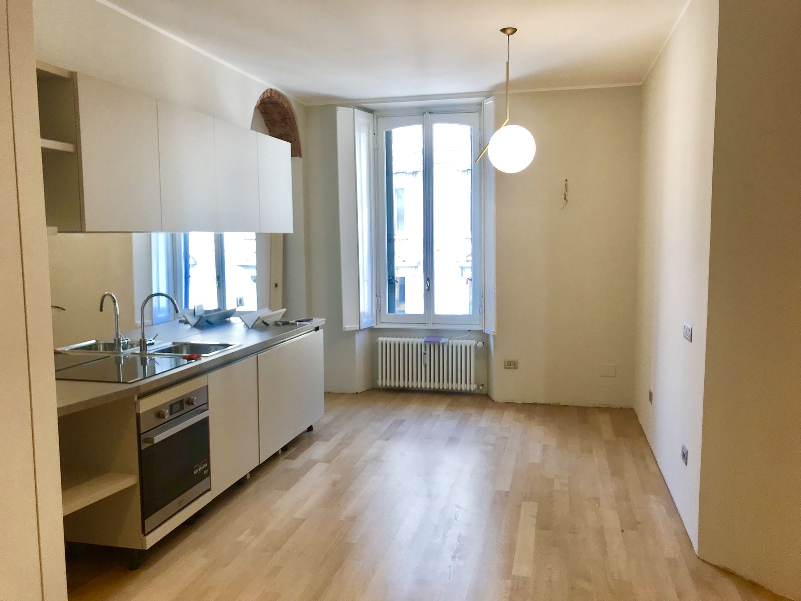 Fotografia della cucina finita: il blocco con piano in acciaio e forno a vista, i pensili bianchi a filo, la fascia a specchio che riflette la finestra e la lampada sferica sospesa