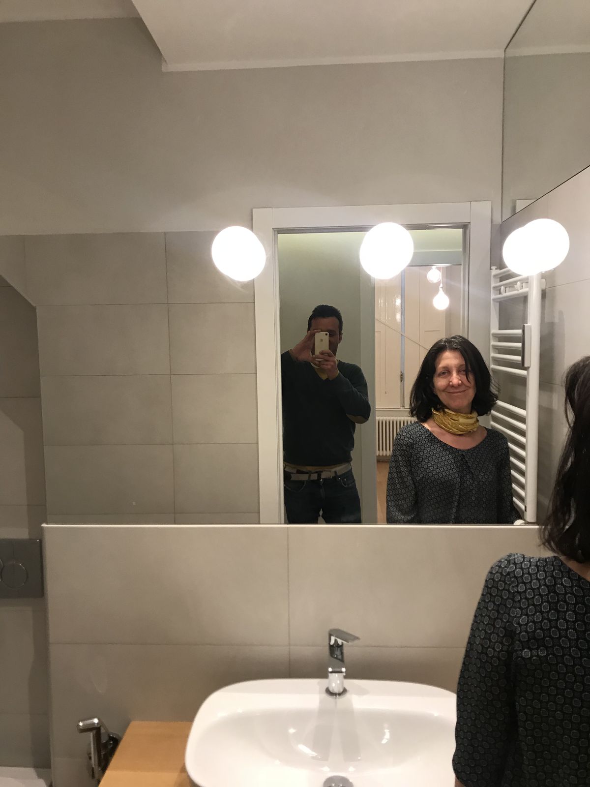Fotografia del bagno cieco finito: lo specchio a tutta parete sopra il muretto piastrellato, tre lampade sferiche Mini-Ball montate sullo specchio, il lavabo e il termoarredo; nel riflesso si vedono due persone