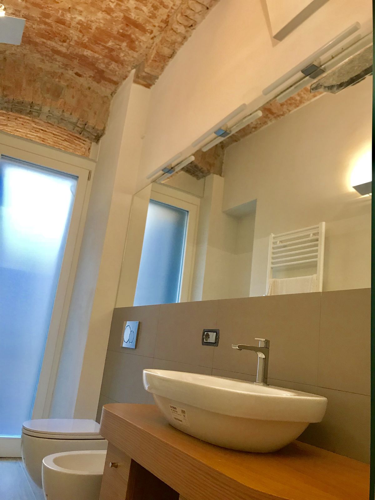 Fotografia di un bagno finito: il lavabo appoggiato sul piano di rovere, lo specchio a tutta parete con la barra di luce, la volta di mattoni a vista e a sinistra un pannello di vetro illuminato