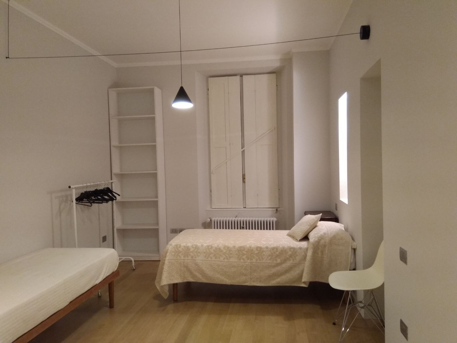 La camera finita: due letti, una libreria alta e stretta, e la lampada a cono appesa a un filo teso che attraversa il soffitto da parete a parete