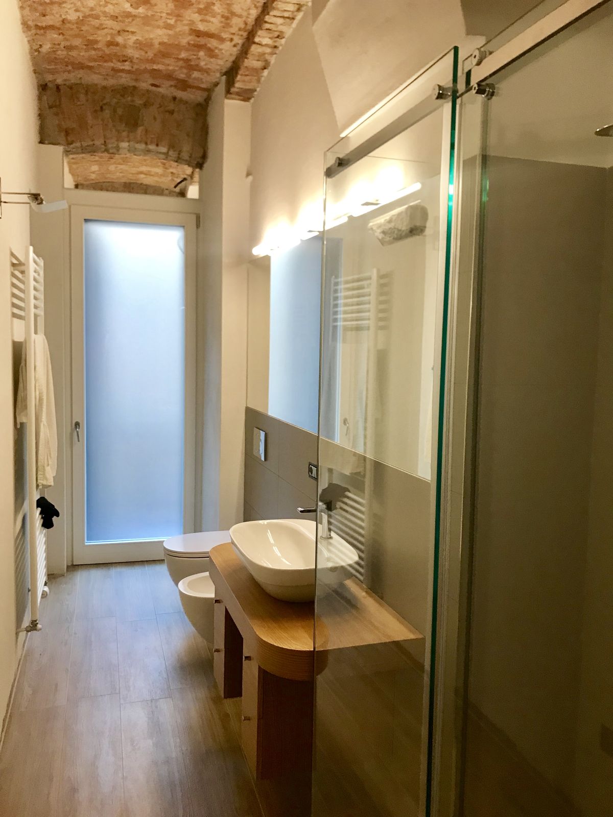 Il bagno padronale fotografato per il lungo: in fondo la finestra a vetro satinato, lungo la parete la consolle di rovere con il lavabo, il vaso e il bidet sospesi, a destra il box doccia in vetro scorrevole, sopra la volta di mattoni a vista