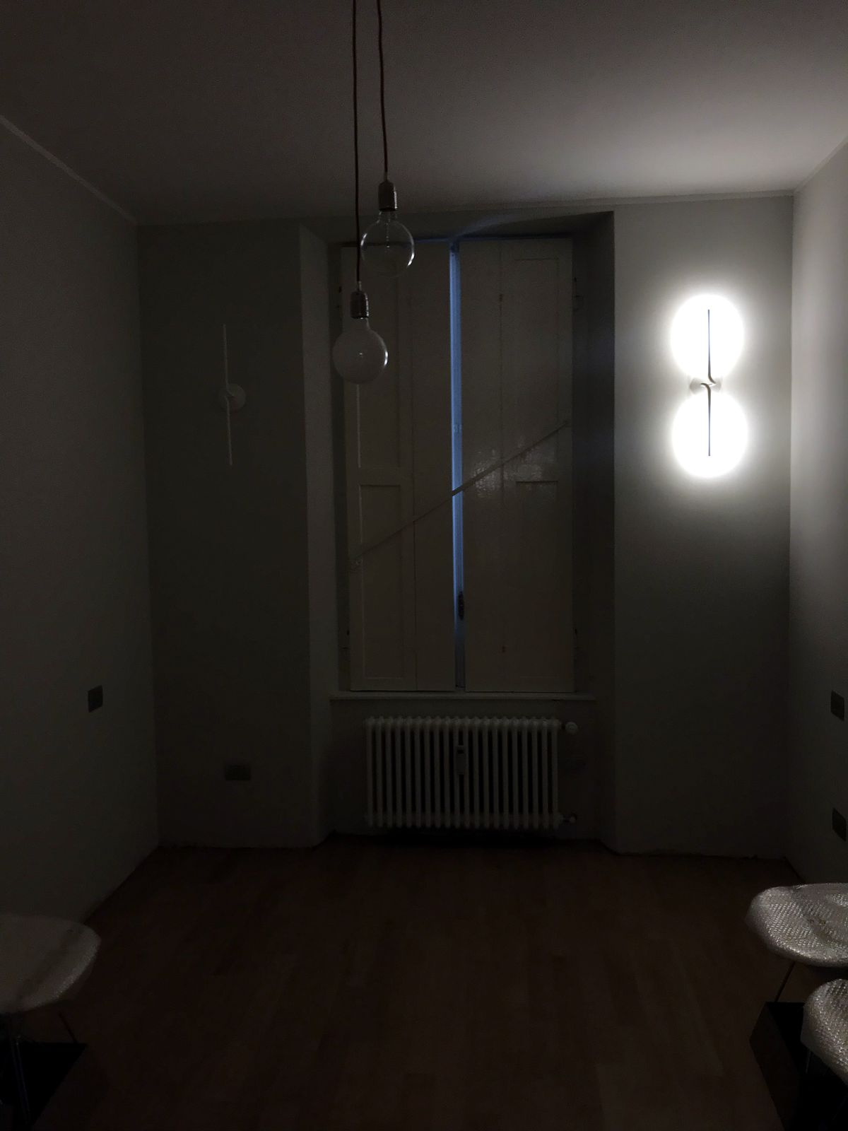 La stanza fotografata al buio: una lampada a sospensione con la lampadina a globo appesa al cavo, e sulla parete di destra un apparecchio a due sfere acceso; in fondo la finestra alta con gli scuri interni chiusi e il radiatore a colonne, in primo piano il bordo di un tavolino e di due sedie