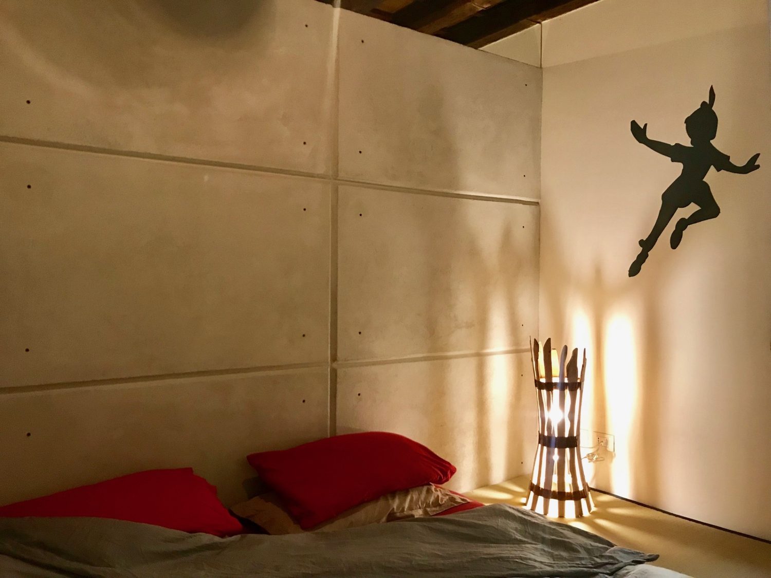 La lampada in ginepro accesa nell'angolo di una camera con la parete in pannelli di cemento, i cuscini rossi e la sagoma di Peter Pan sul muro