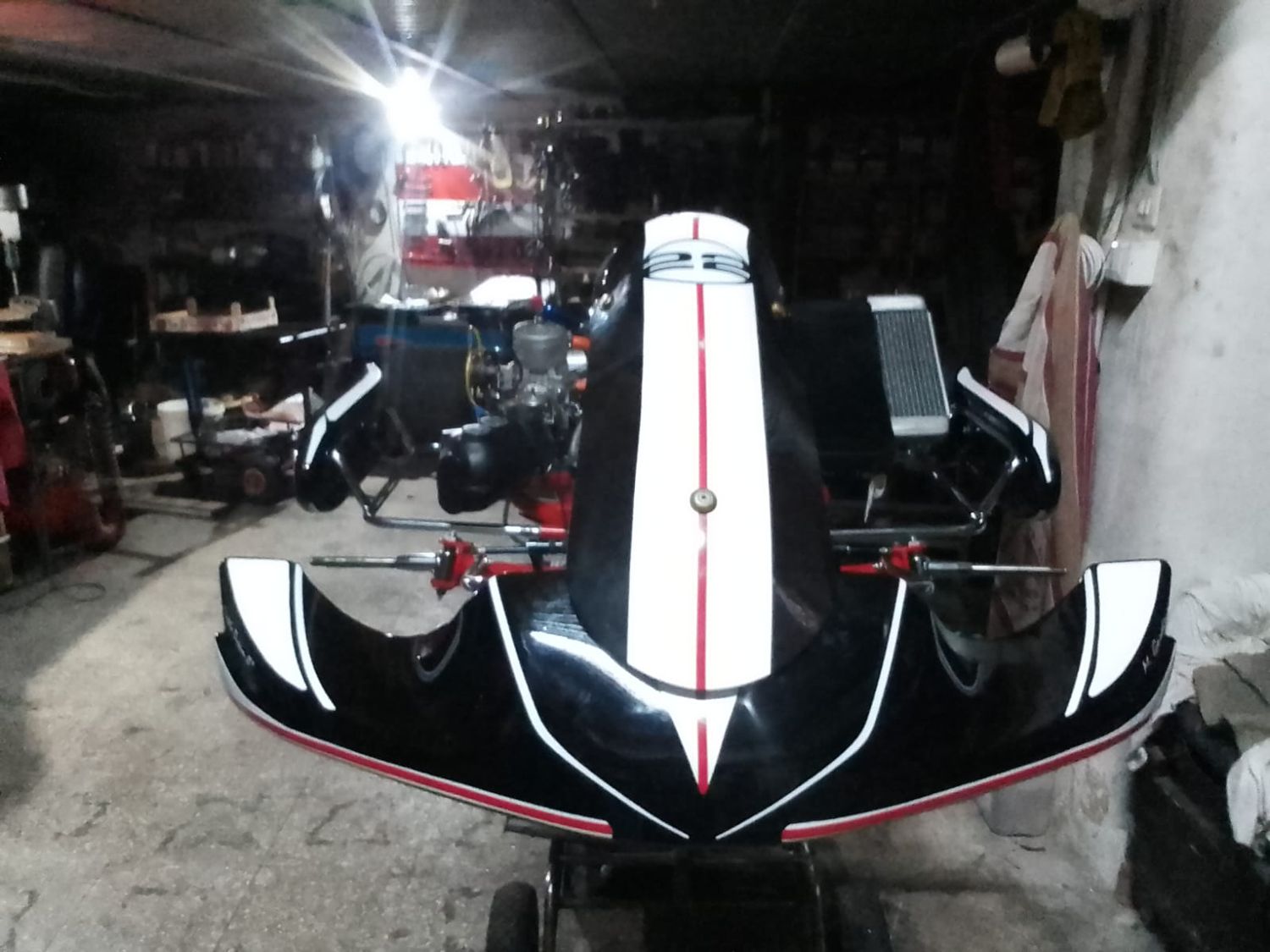 Il muso del kart finito: carena nera e bianca lucida con una riga rossa che scende al centro, nella penombra dell'officina