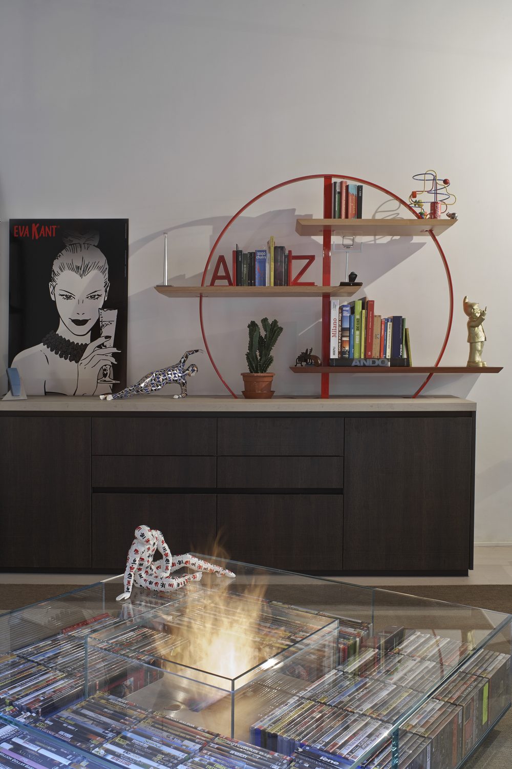 La libreria: un cerchio di piatto d'acciaio rosso che esce dal top del mobile scuro, con tre mensole di legno diverso al suo interno, libri e oggetti