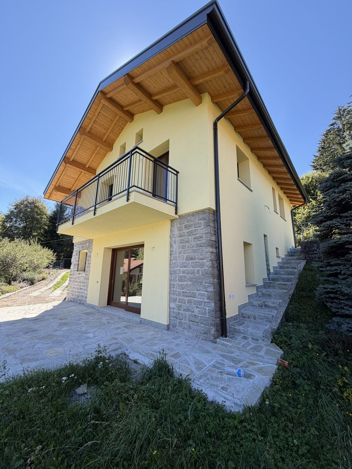 La casa finita: il volume intonacato di giallo chiaro, il basamento in pietra, il tetto in legno a vista e il balcone