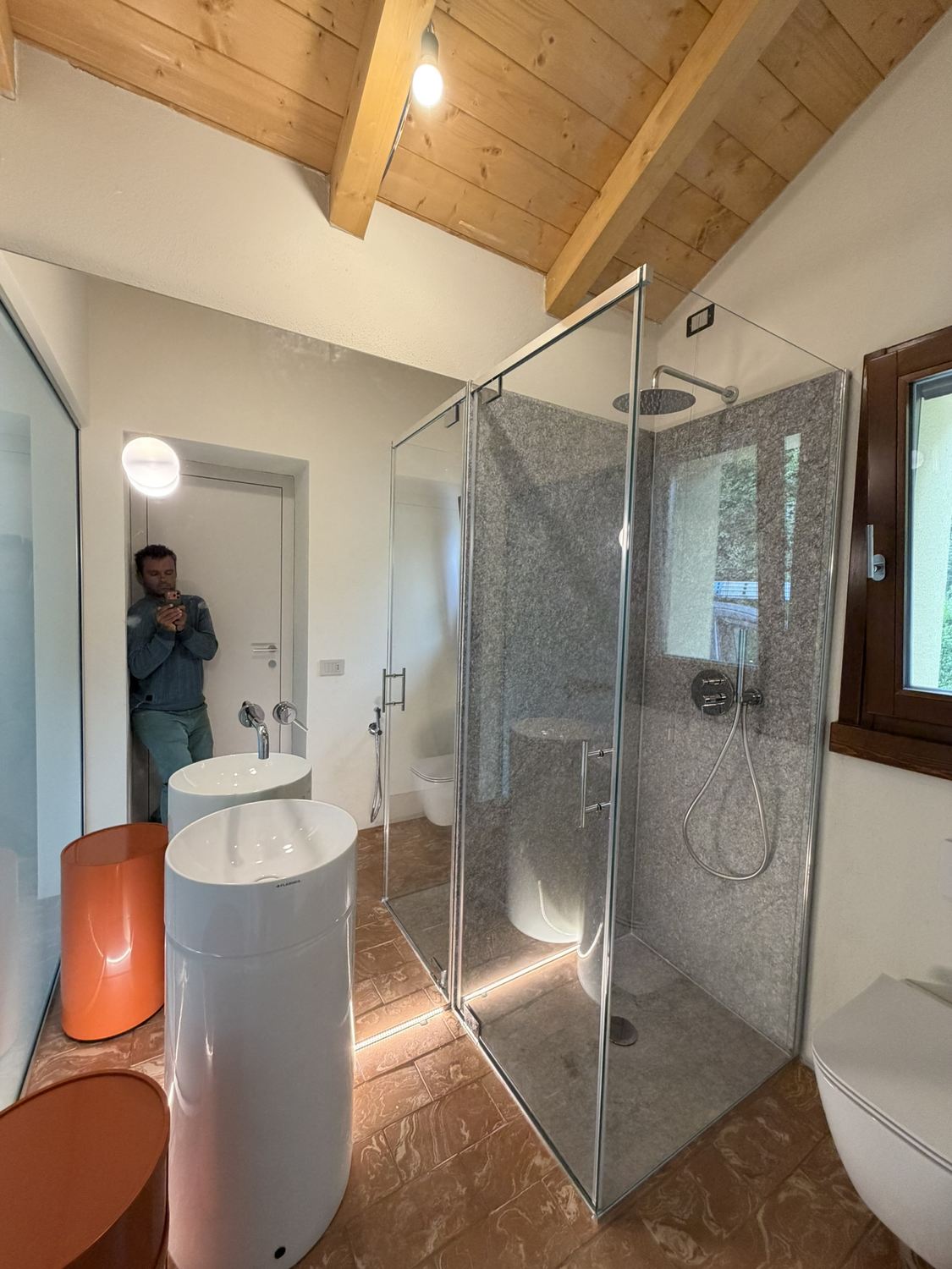 Settembre 2026: lo stesso bagno finito, con la doccia in vetro, il lavabo cilindrico, il pavimento in cotto e le travi del tetto a vista