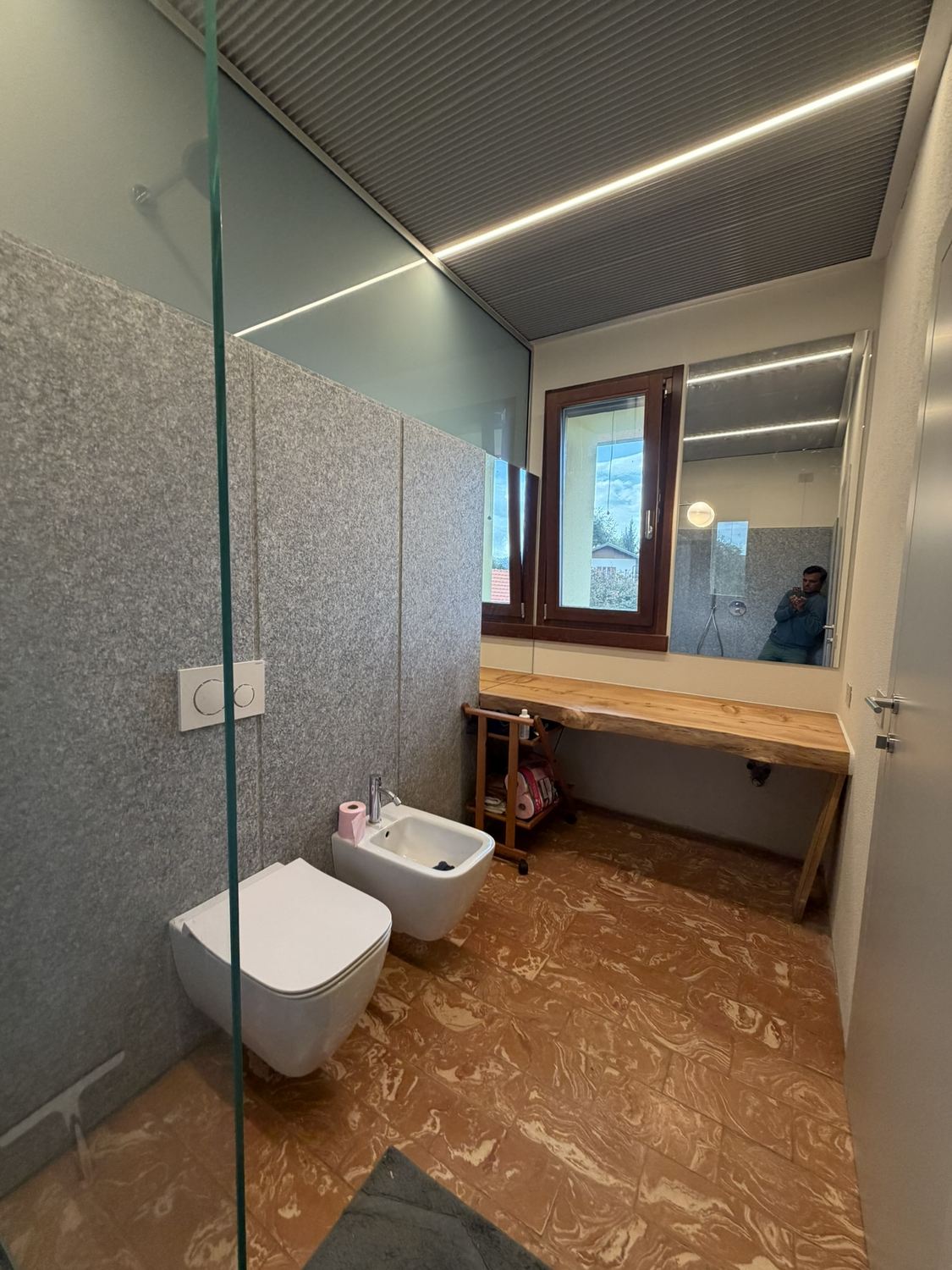 Il bagno principale ripreso da dentro la doccia, oltre lo spigolo della lastra di vetro: a sinistra la parete di pietra, al centro vaso e bidet sospesi, a destra la finestra con lo specchio a fascia e sotto un piano di legno massello col bordo a spacco