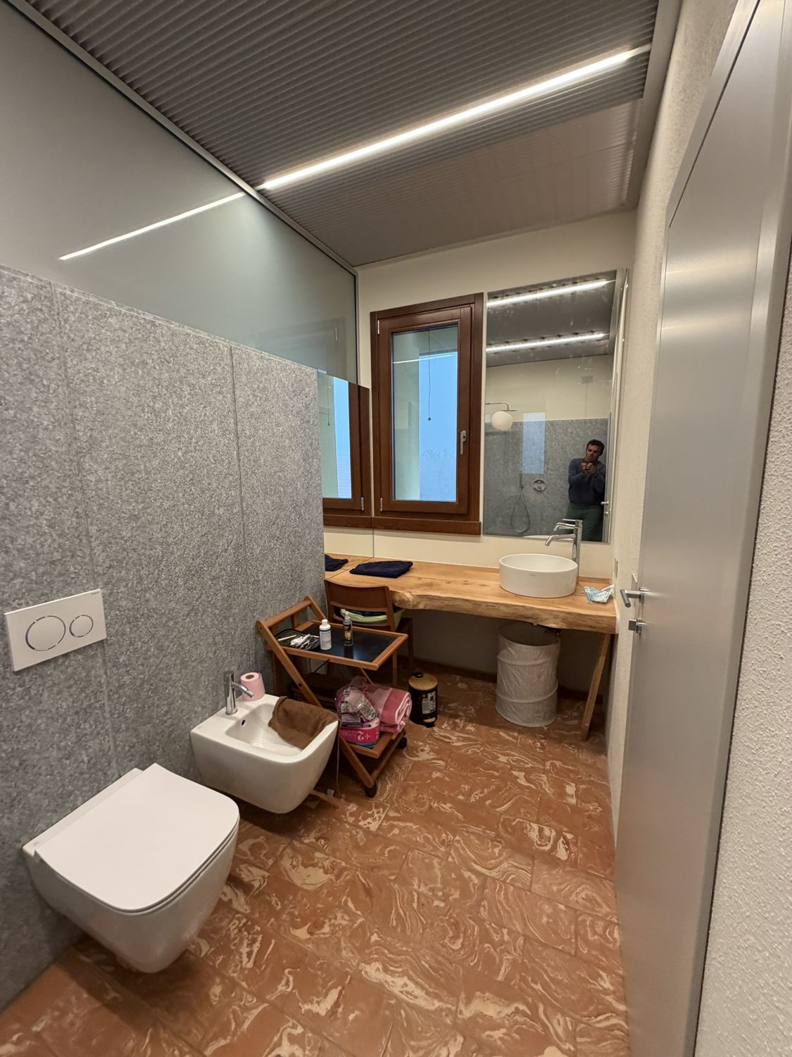 Lo stesso bagno verso la finestra: sul piano di legno massello il lavabo tondo Laufen con il miscelatore alto, lo specchio a fascia con la luce led, le pareti in pietra grigia e il pavimento in cotto