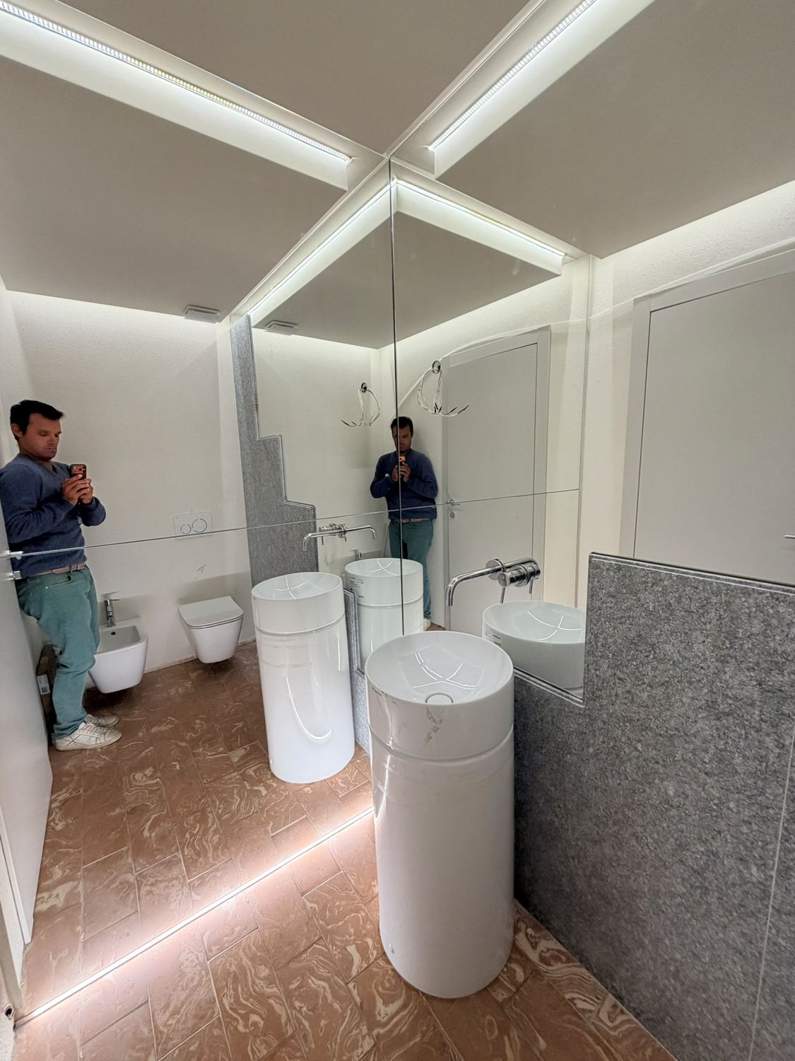 Il bagno a specchi montati: lavabo cilindrico a colonna con rubinetto a muro, rivestimento in pietra grigia che si interrompe con un bordo a gradini e riparte nel riflesso, strisce LED a soffitto e a pavimento, bidet e wc sospesi sul fondo, e l&rsquo;architetto riflesso mentre fotografa