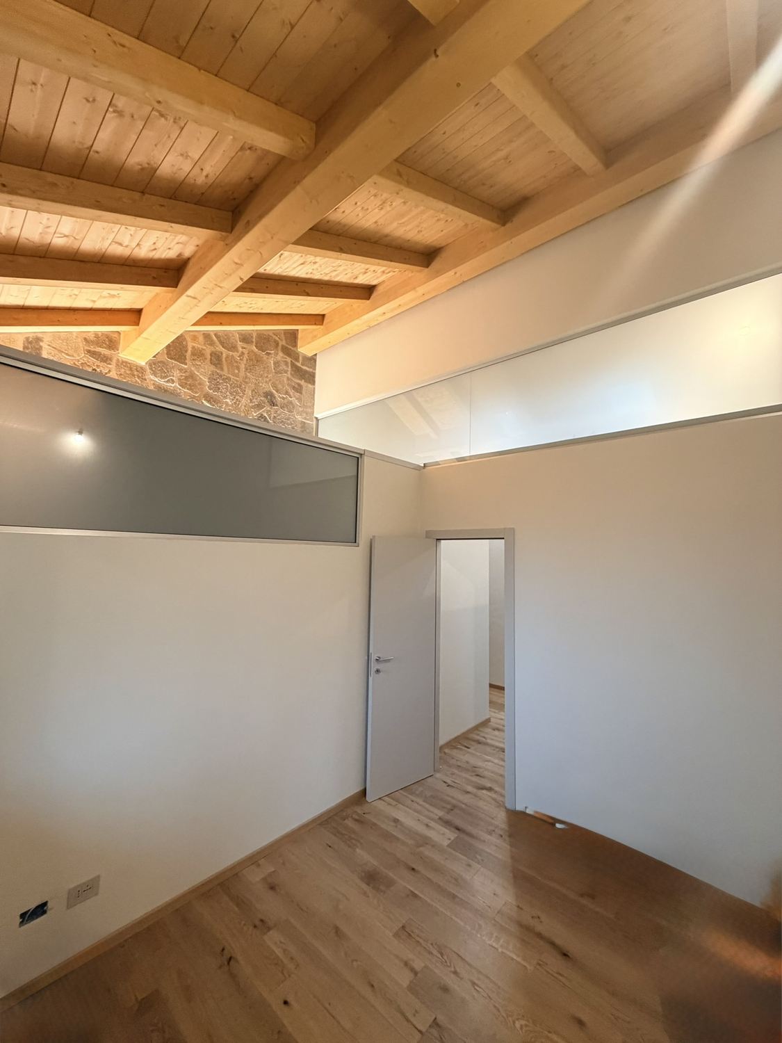 La camera degli ospiti a luci accese: pavimento in rovere, pareti chiare, la striscia di vetro accoppiato bianco latte sopra il muro basso e, oltre, il vecchio muro in pietra del nevaio illuminato dal basso, sotto le travi nuove del tetto