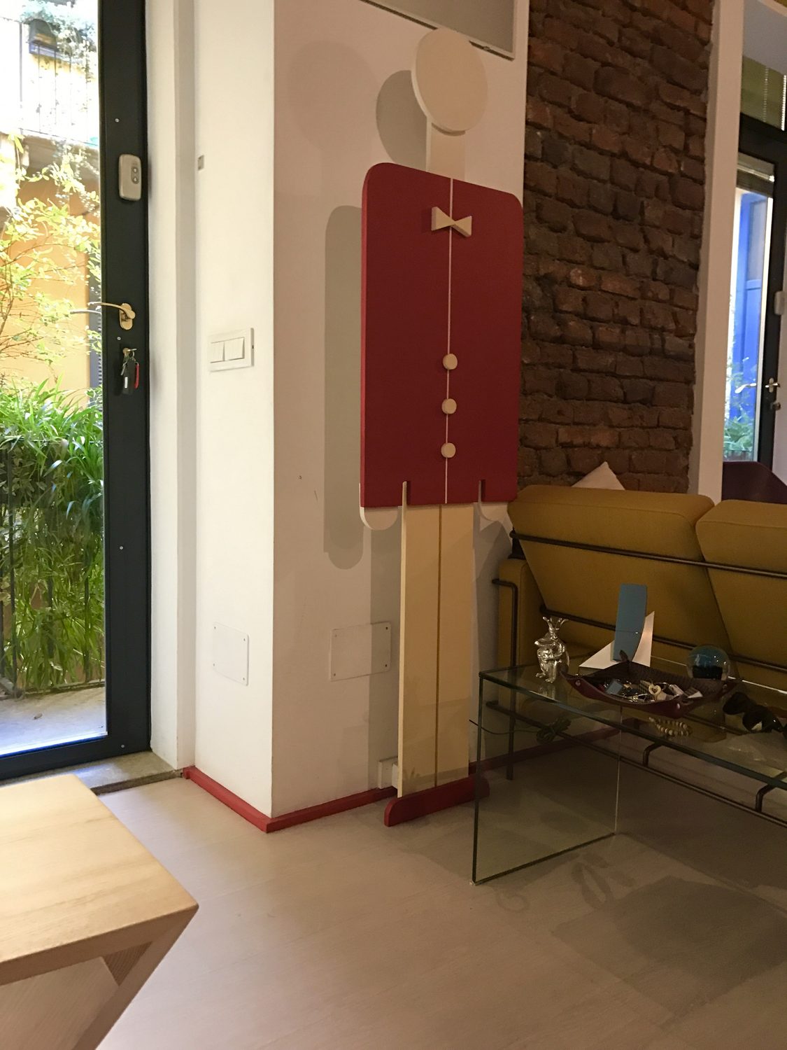 L'Omino Appendino appeso al muro accanto a una porta finestra: giacca rossa con papillon e tre bottoni bianchi, testa tonda, pantaloni chiari