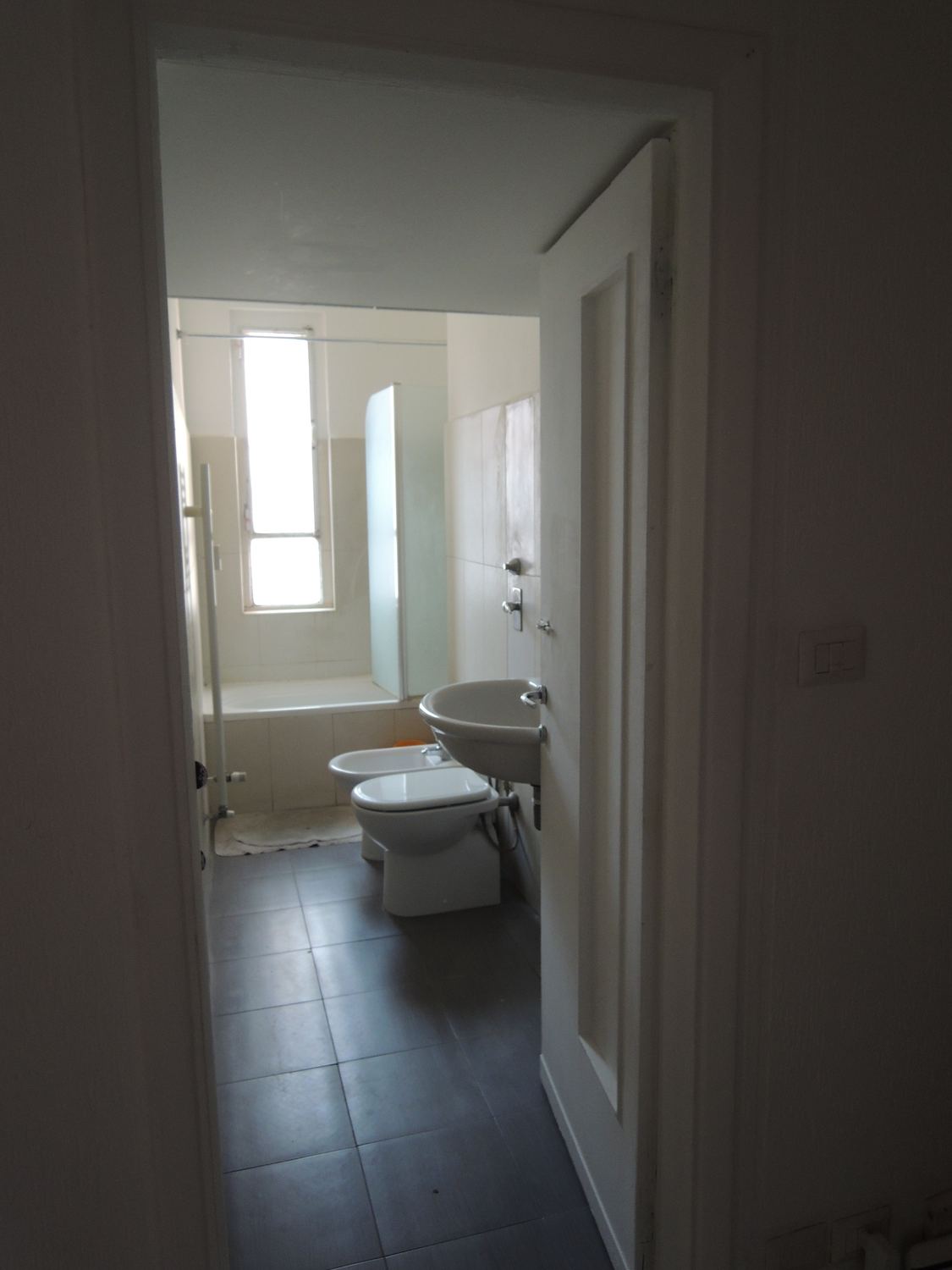 Il bagno prima, visto dalla porta: lungo e stretto, pavimento scuro, sanitari in fila contro la parete