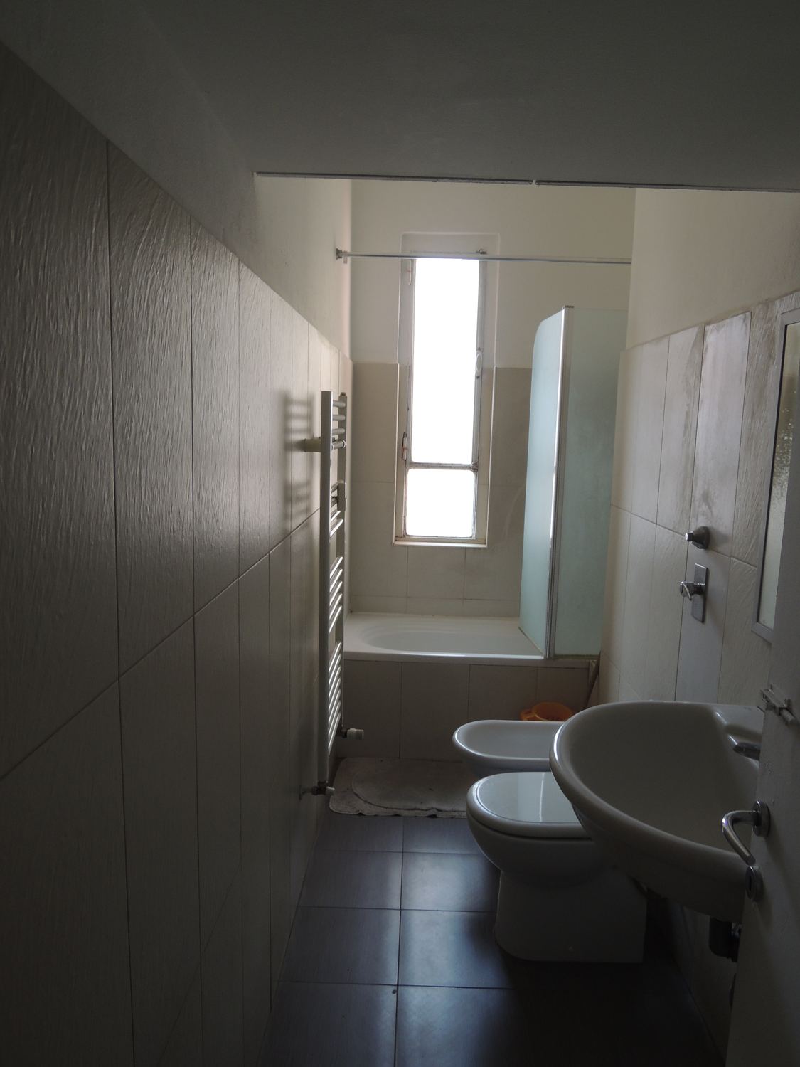 Il bagno prima: stretto, piastrelle chiare, vasca e sanitari in fila, la finestrella in fondo