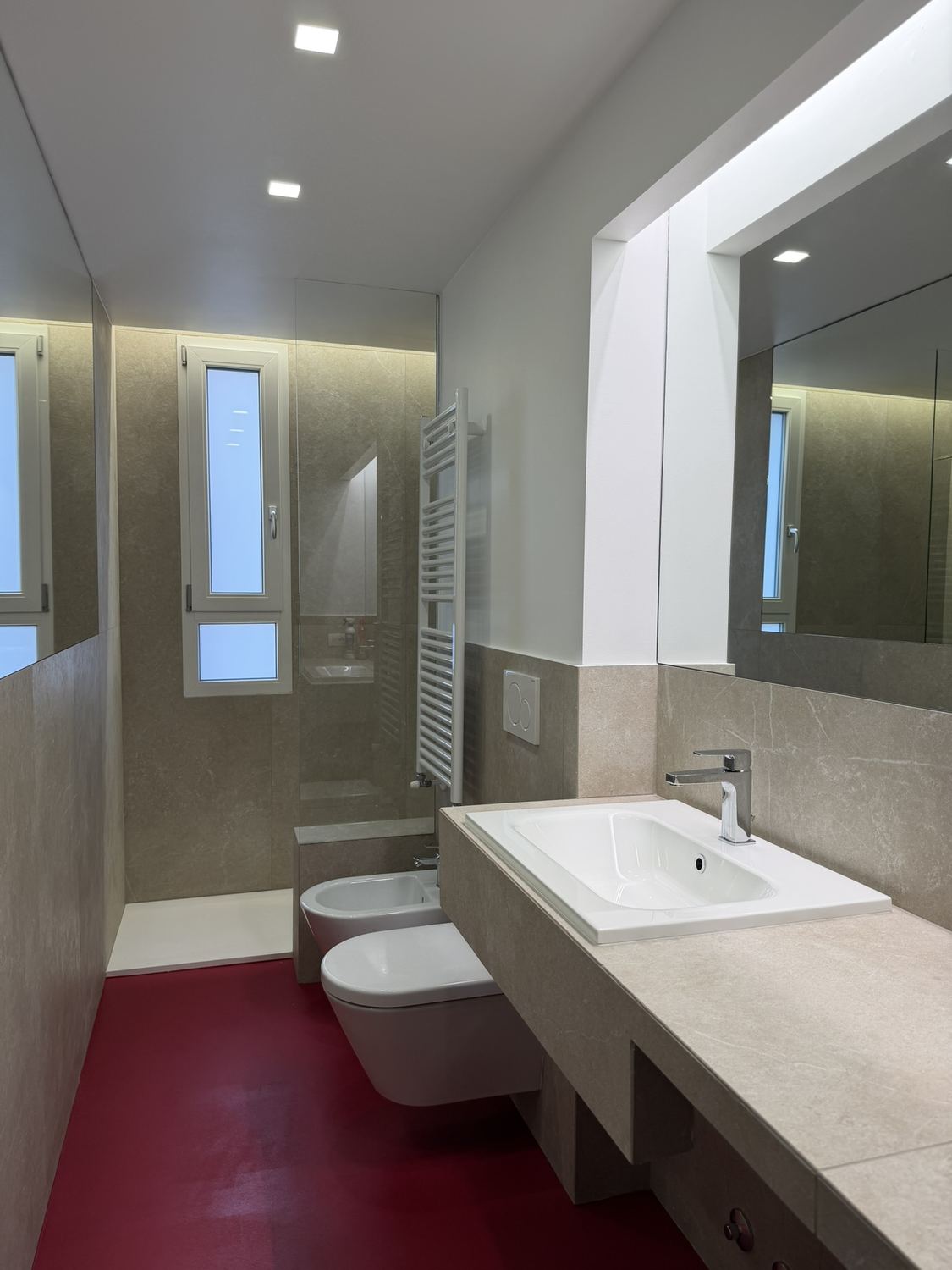 Il bagno finito visto dalla porta: la lunghezza intera, il pavimento rosso, il piano del lavabo a sbalzo e lo specchio illuminato