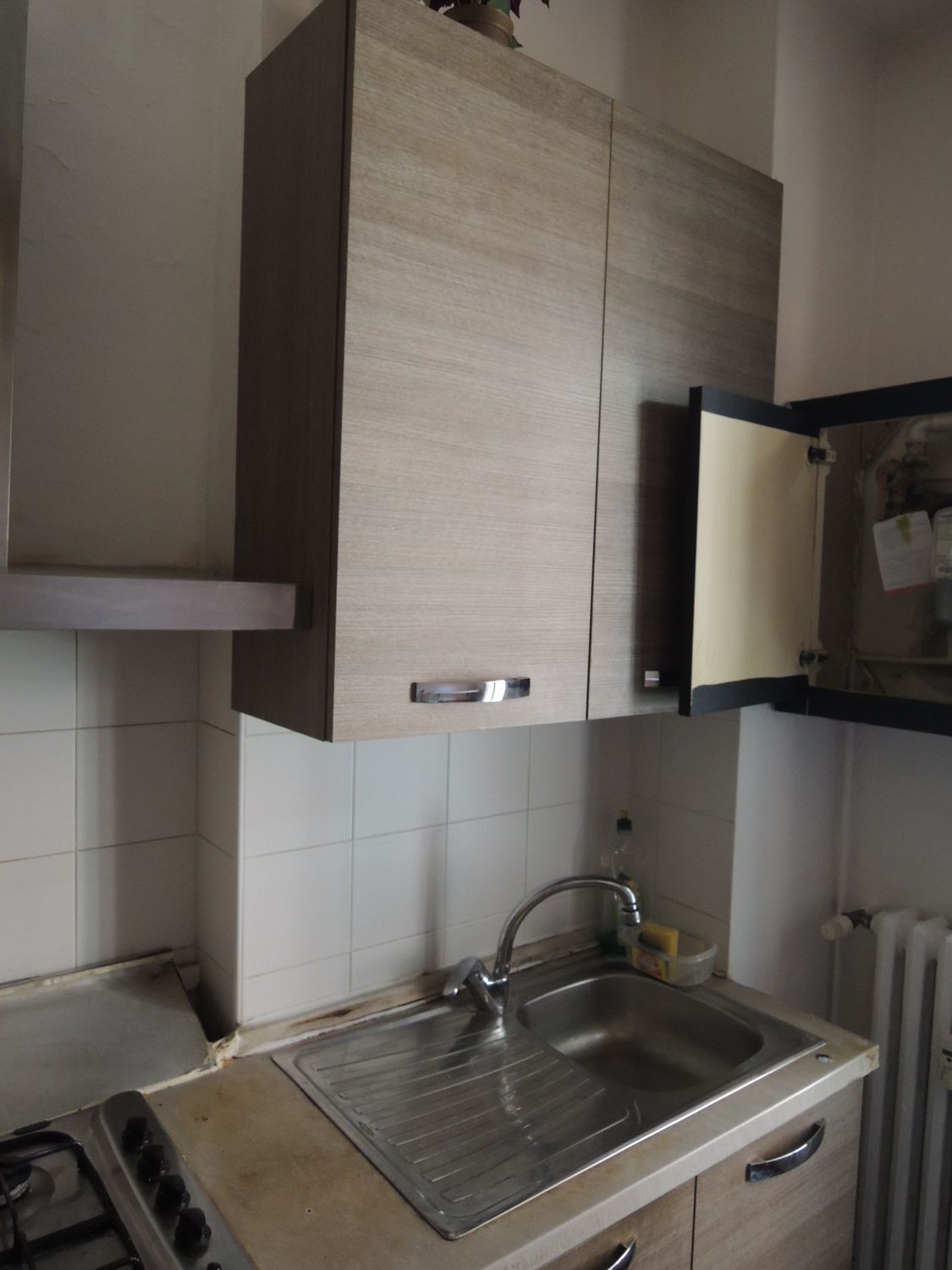 La vecchia cucina: pensili in laminato, lavello sotto la finestra, piastrelle e radiatore