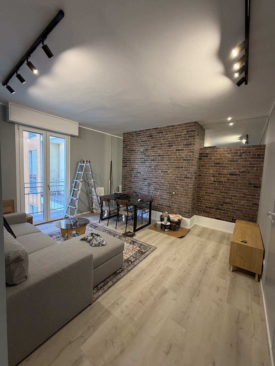 Il soggiorno: la parete alta di mattone antico, a destra il muro di mattone pi&ugrave; basso e sopra di esso la fascia a specchio che riflette i faretti del soffitto