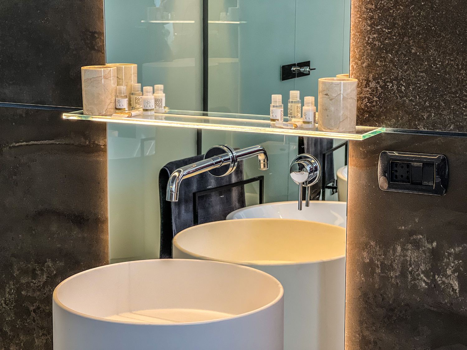 Lavabo cilindrico e mensola in vetro illuminata dall'interno contro la parete in pietra