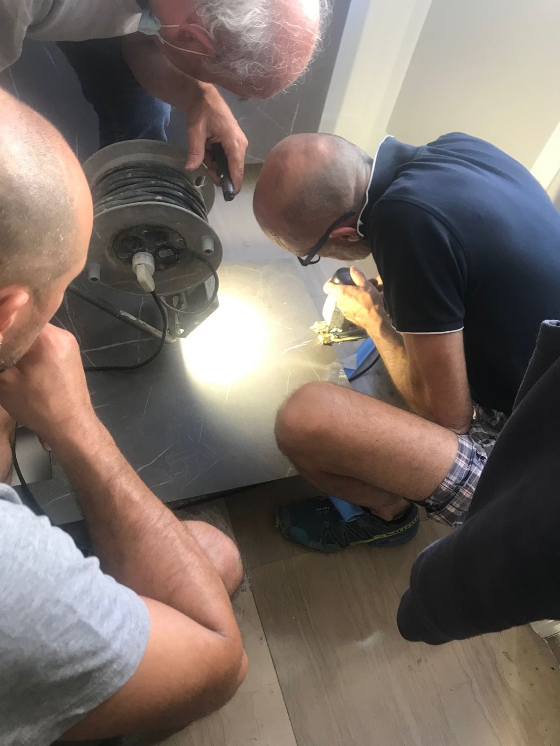 Tre uomini accovacciati per terra intorno a una lastra di pietra scura, con una lampada da cantiere che la illumina di taglio