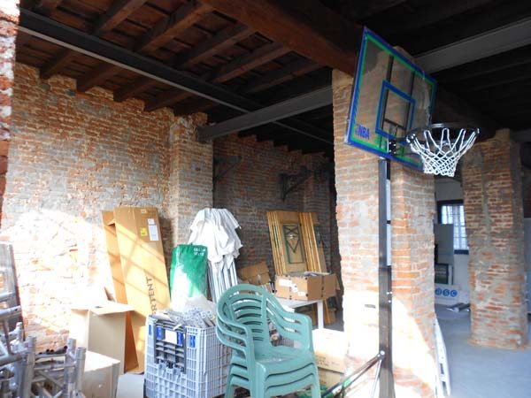 Il locale come deposito: sedie di plastica impilate, scatoloni, un canestro da basket sul pilastro di mattoni